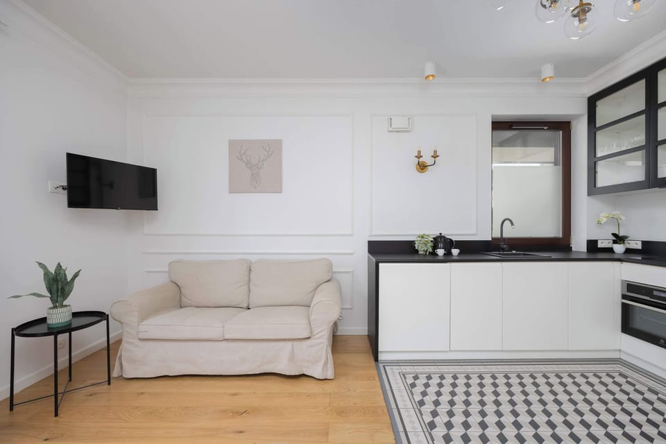 A living room with a wall-mounted TV, white couch, and a black-and-white tiled floor, connected to a kitchenette.

