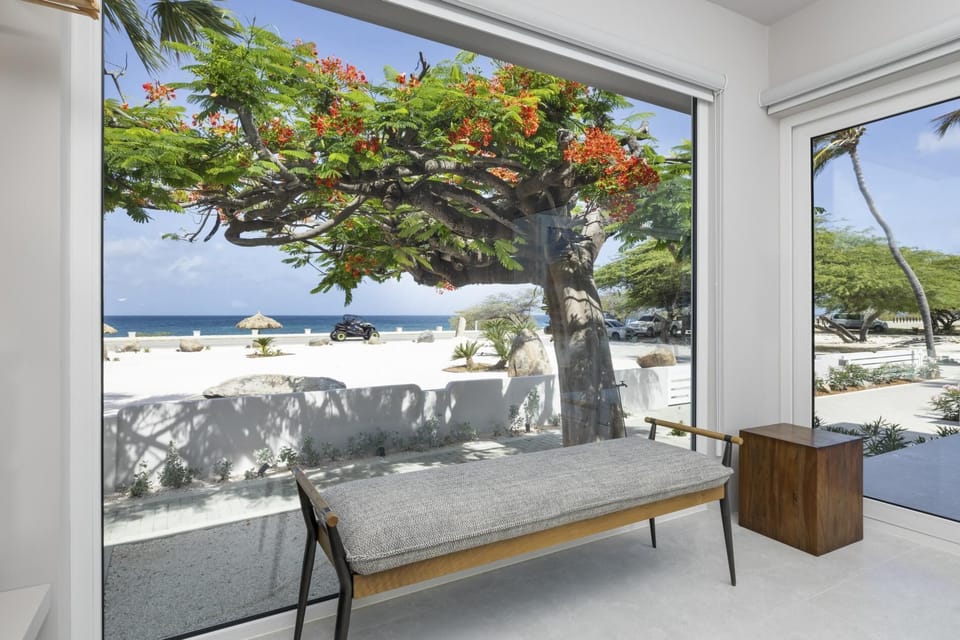 Cozy indoor bench framed by floor-to-ceiling windows, with lush blooms and ocean views just steps from your door—ideal for morning coffee or quiet reading.