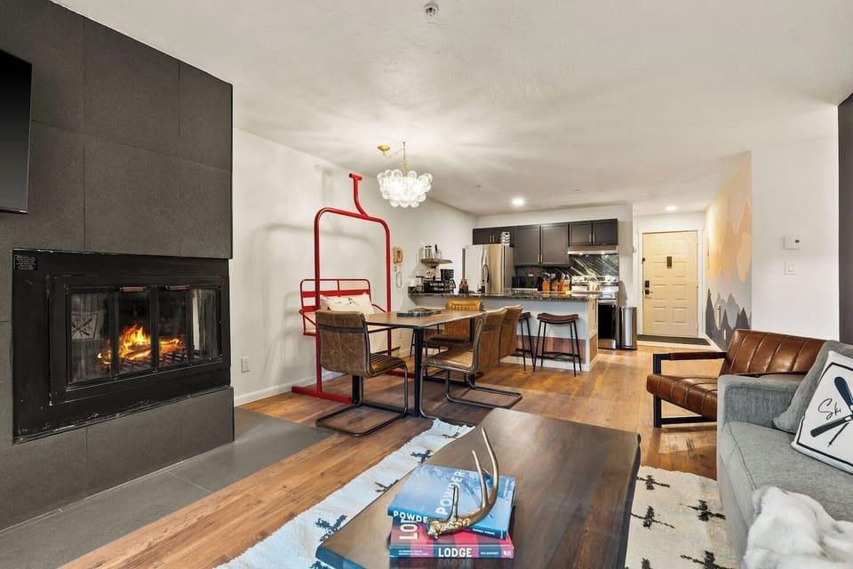 Grab a book off the coffee table stack and claim a seat by the fire — from here you can see the table set for dinner and the kitchen still going in the background.