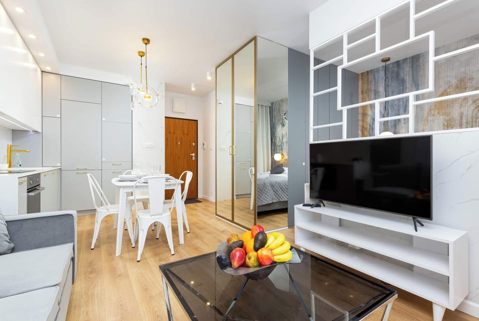 An open-concept layout combining a living room, kitchen, and dining area. The room has stylish white furniture and large windows, giving the space an airy feel.