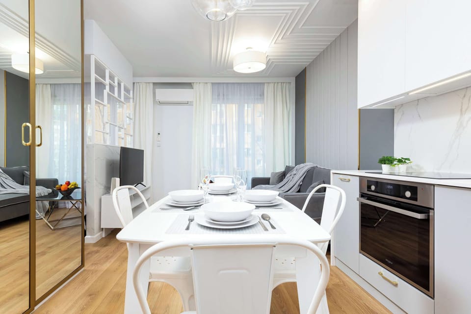 A modern, open kitchen with a white dining table placed nearby. The area is bright and airy, with white furnishings and gold accents.