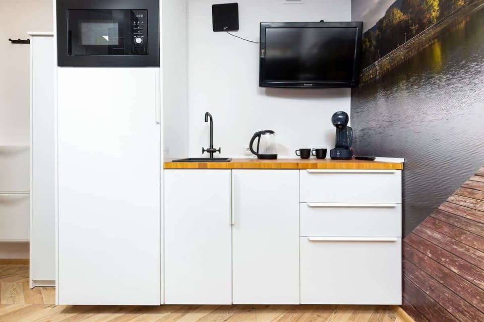 A compact kitchenette with white cabinets, a small sink, and a TV mounted above — no window in sight.