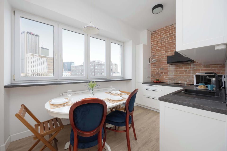 A modern kitchen with a round wooden dining table and navy chairs, next to large corner windows. Brick accents add warmth to the space.