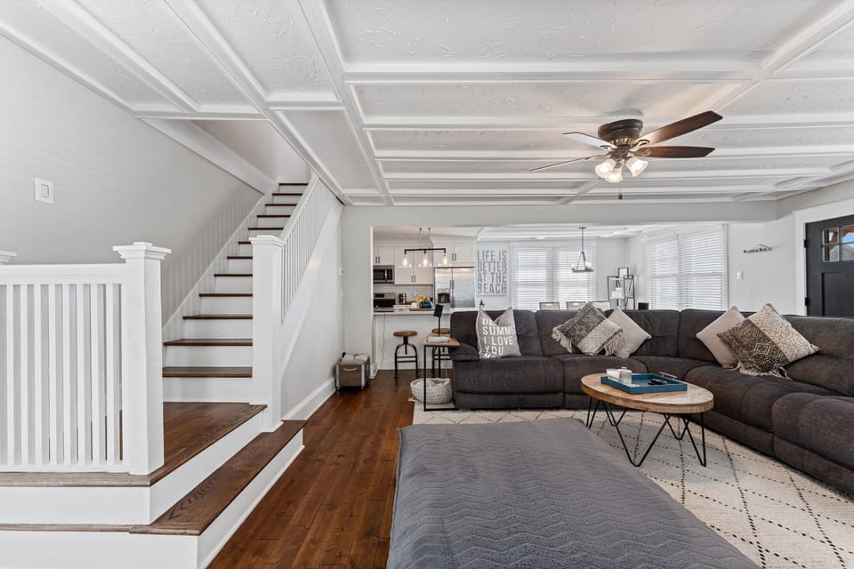 Bright, modern living room with a dark gray sectional, round coffee table, and textured rug over hardwood floors. A white coffered ceiling, coastal décor, ceiling fan, and staircase add a relaxed yet refined feel.