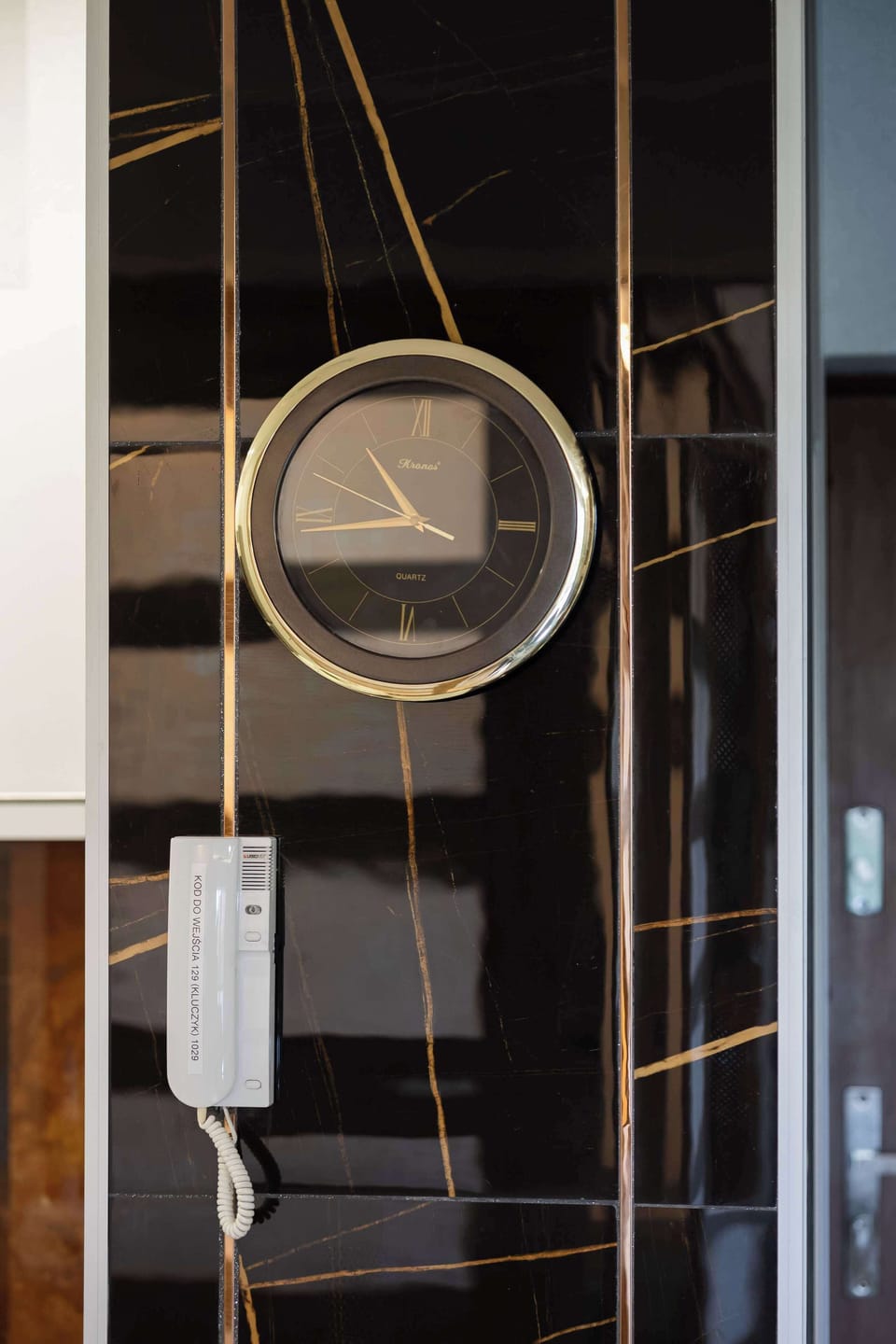 A hanging wall clock with metallic trim and a leather-style strap, placed against a glossy dark wall.