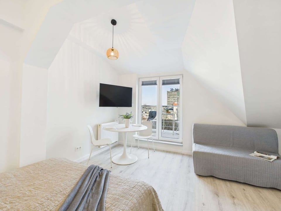 A modern living room area with a gray couch and TV. Glass doors lead to a balcony, filling the room with daylight.