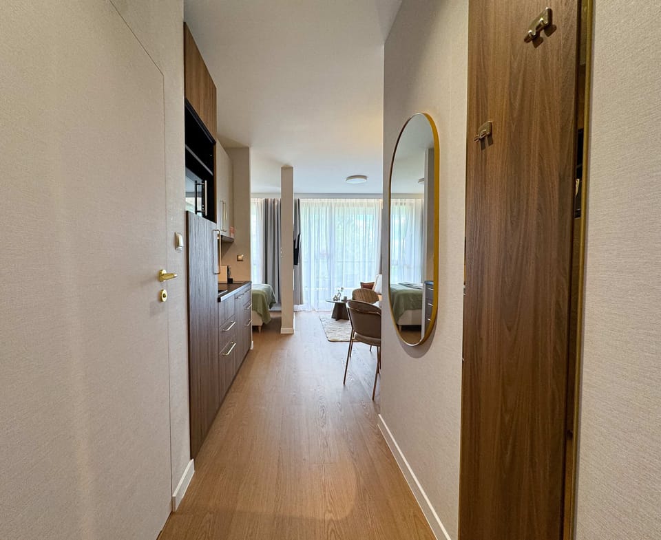 A long, bright corridor leading to the living room and bedroom, featuring wooden flooring and white walls adorned with tasteful paintings.