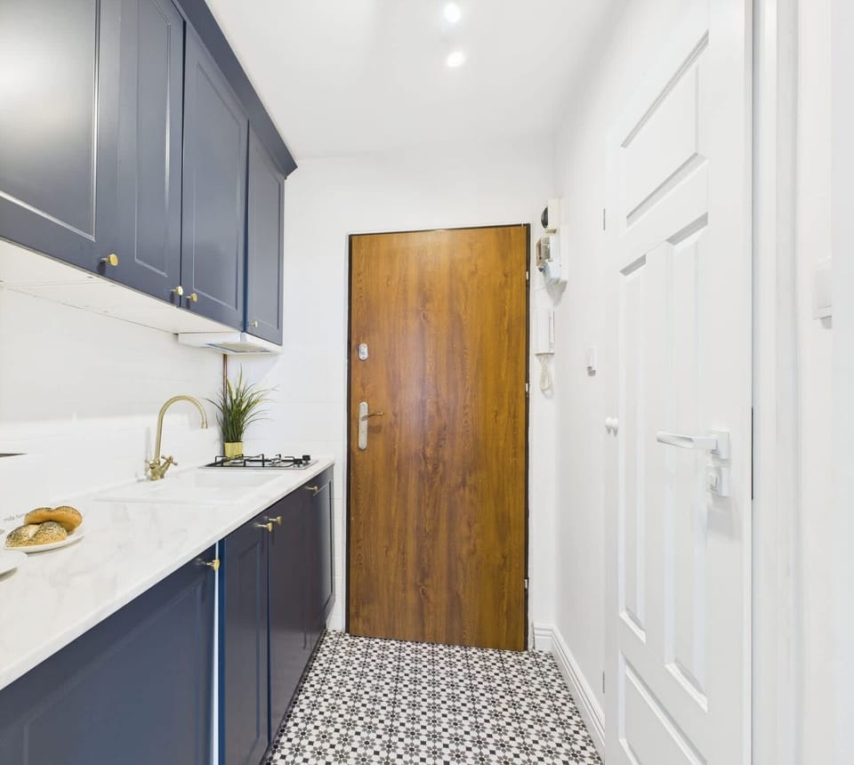 A narrow entryway kitchen featuring deep blue cabinets, a gold faucet, patterned floor tiles, and a warm wooden front door creating a welcoming contrast.