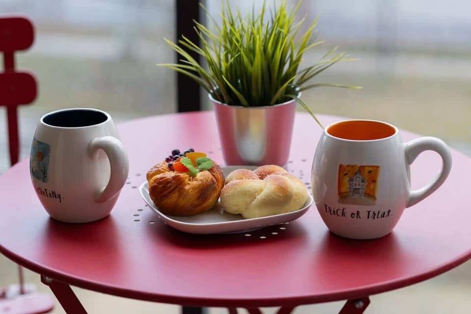 A cozy breakfast setup on a red balcony table, featuring two cups of coffee, a plate with fresh pastries, and a small green plant, creating a relaxing atmosphere.
