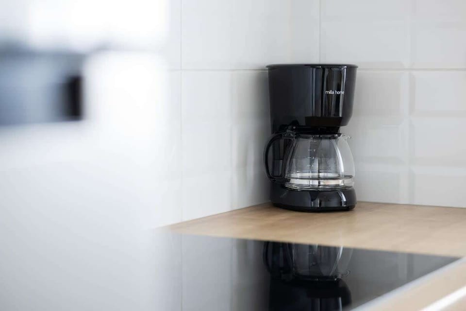 A compact kitchen corner featuring a black drip coffee machine on a small countertop. White tiles provide a simple, clean backdrop.
