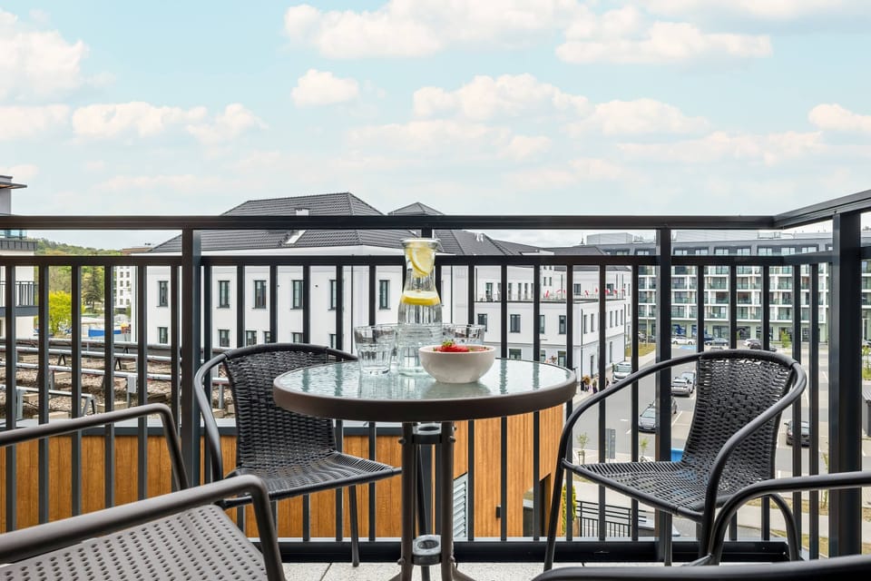 A balcony with a round table and four chairs invites you to relax - on the table a pitcher of lemonade, glasses and a bowl of strawberries create a summer atmosphere with a view of modern buildings.