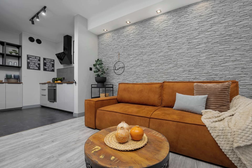 Another angle of the living room with the orange sofa, showcasing the open layout leading into the kitchen. The design combines warm tones with industrial elements.