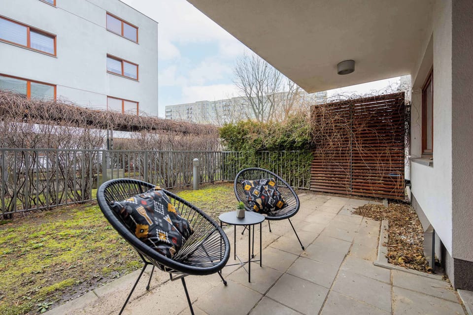 A small outdoor patio area with two black chairs and a round table. The space is surrounded by a low fence and greenery.