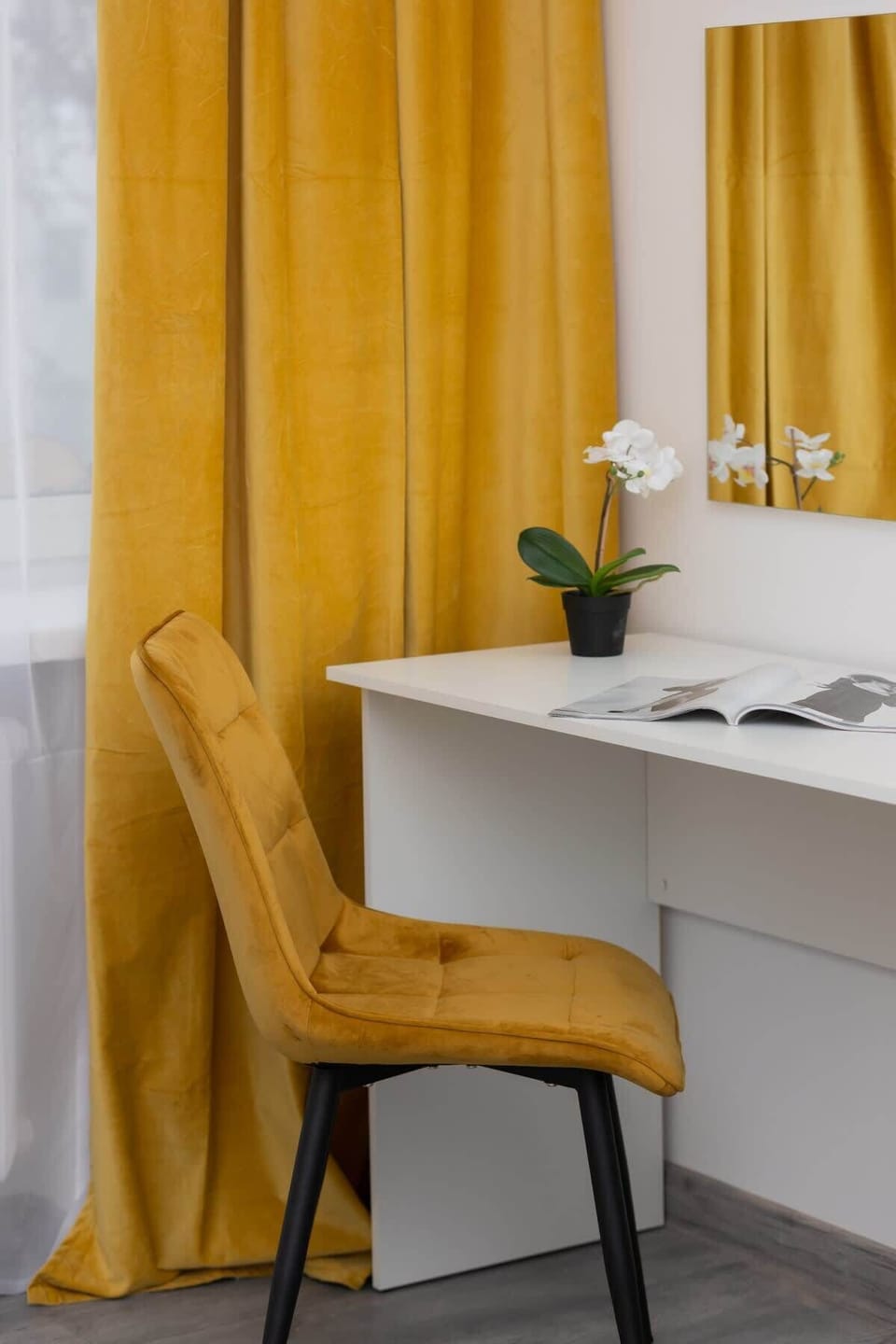 A stylish bedroom corner with a white desk and mustard curtains. The decor is simple and elegant, creating a cozy workspace.