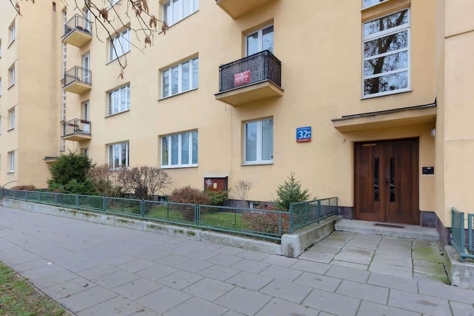The exterior view of the apartment building with yellow walls and balconies. A small green area surrounds the entrance.