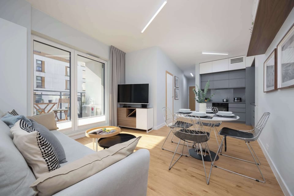 A modern open-plan living room with a sofa, dining table, and chairs. The decor is bright and minimalistic, featuring wooden flooring.