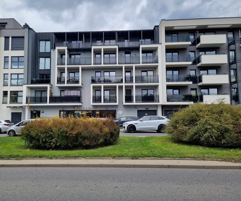  A modern apartment building with multiple balconies, surrounded by greenery and bushes in front. The building has a contemporary design with large windows and white and grey accents.