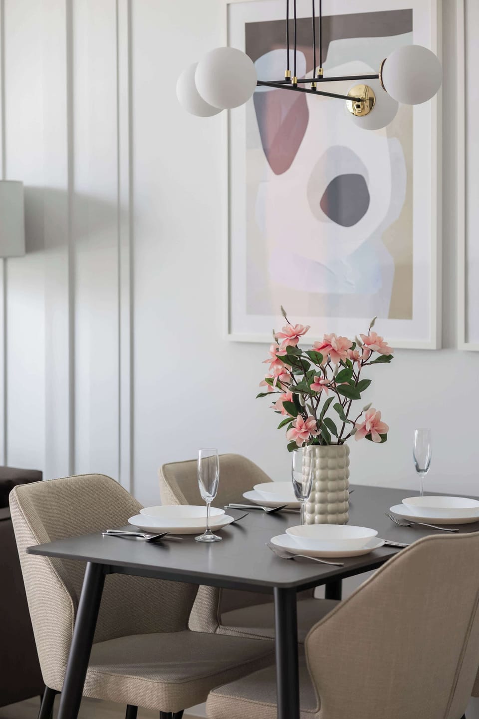 A stylish dining area in a modern studio with a dark table set for four, surrounded by beige upholstered chairs, decorated with an elegant bouquet of pink flowers and illuminated by a designer lamp with spherical bulbs and gold accents.