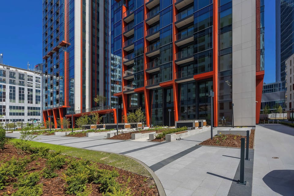 A modern high-rise building with a glass facade and red structural elements, surrounded by greenery and set against a clear blue sky, offers a prestigious location for a comfortable studio.