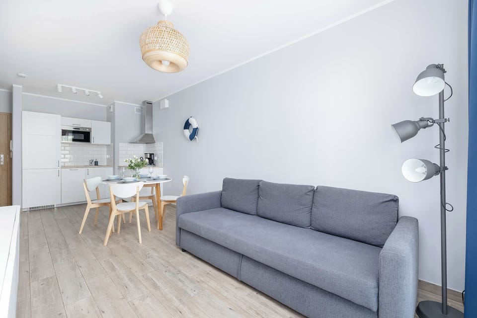 Bright and comfortable living room with grey double sofa bed, dining table with chairs, and kitchenette.
