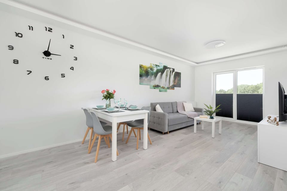 A spacious living and dining area with light wooden flooring, a modern clock on the wall, and a table set for four.