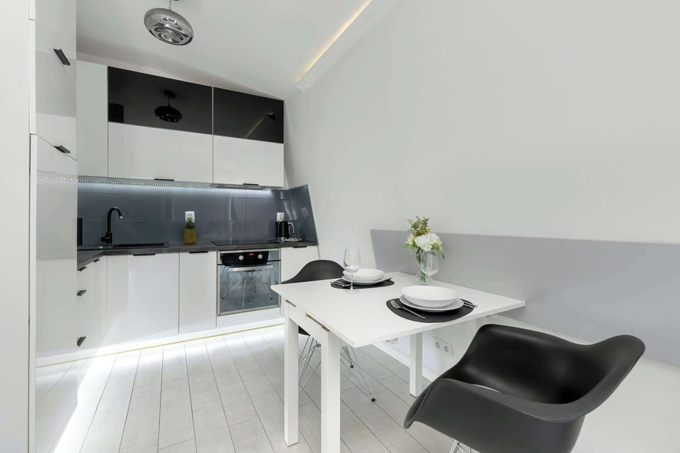 A minimalist dining space with a small black table, two modern chairs, and a built-in kitchenette with white cabinetry.

