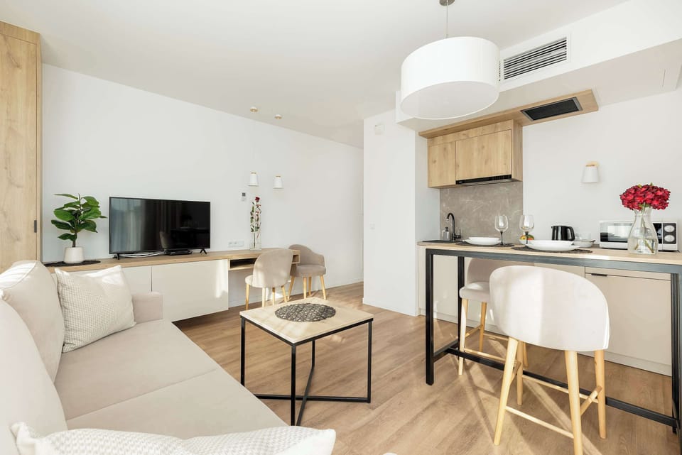  This open-plan living room features a light sofa and a small dining table in front of a modern kitchen. A white pendant lamp hangs above, and the large windows create a bright and airy feel.