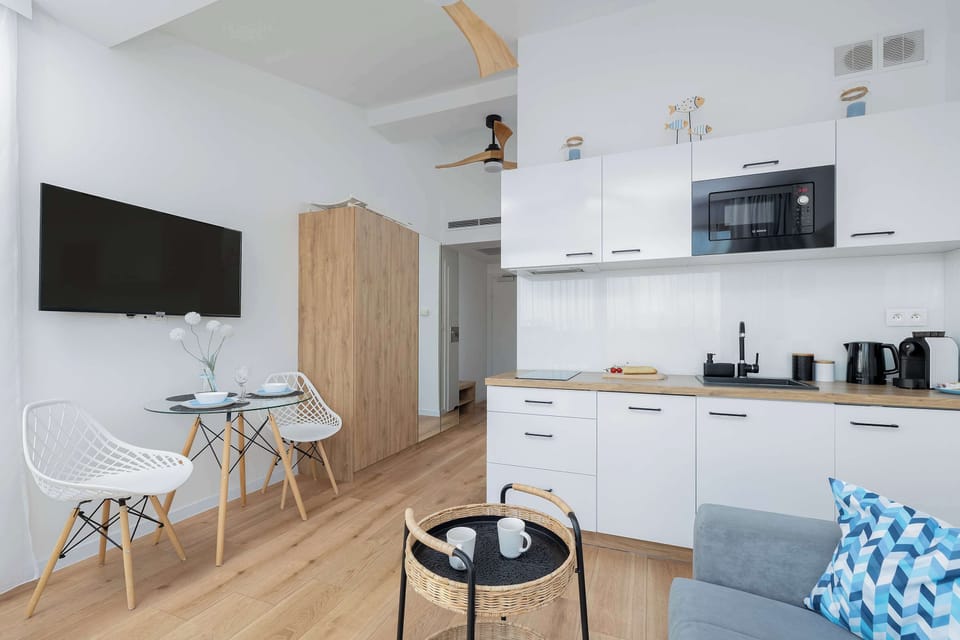 A bright living room with a small dining area, wooden flooring, and a TV mounted on the wall.