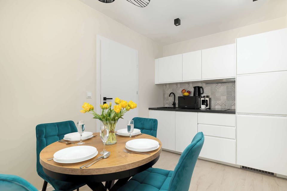 A small dining nook with a round table and two teal chairs, placed near the kitchen. Yellow flowers add a fresh touch to the minimalist interior.