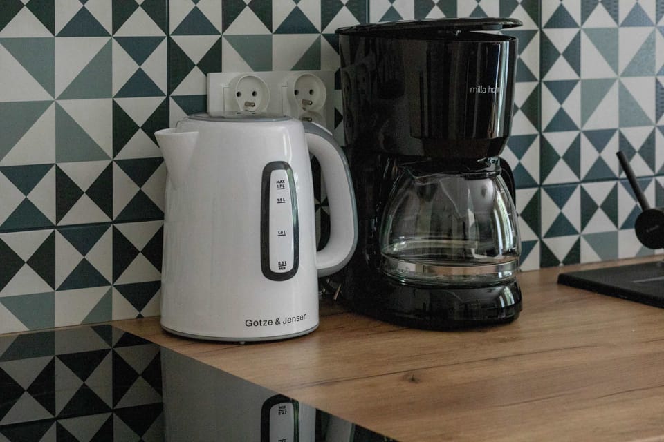 A close-up of a countertop with a white electric kettle and black coffee machine. The background features a trendy geometric-patterned backsplash.