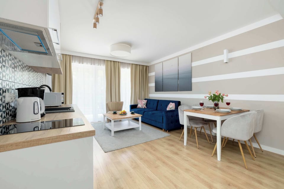 A living area with a navy corner sofa, white coffee table, and a large window that brings in natural light. A kitchen can be seen in the background, forming part of an open-concept space.