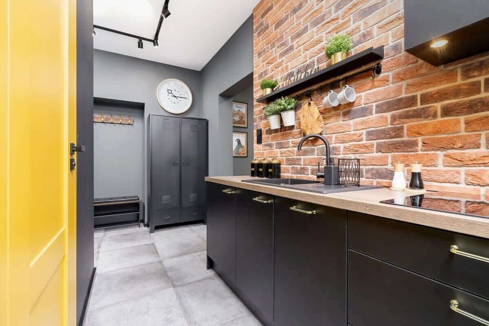 A compact kitchen with brick walls, black cabinetry, and yellow highlights. Industrial-inspired lighting and a mix of modern and rustic elements stand out.
