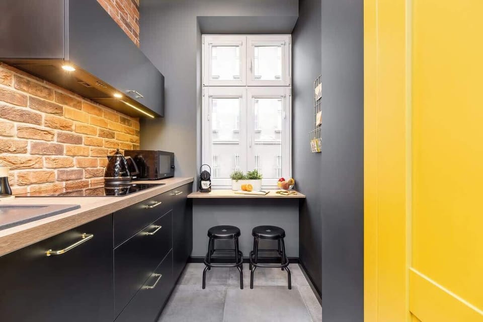 A well-lit kitchen with a large window, brick accent walls, and modern black and yellow decor. Bar stools are neatly placed under the countertop.