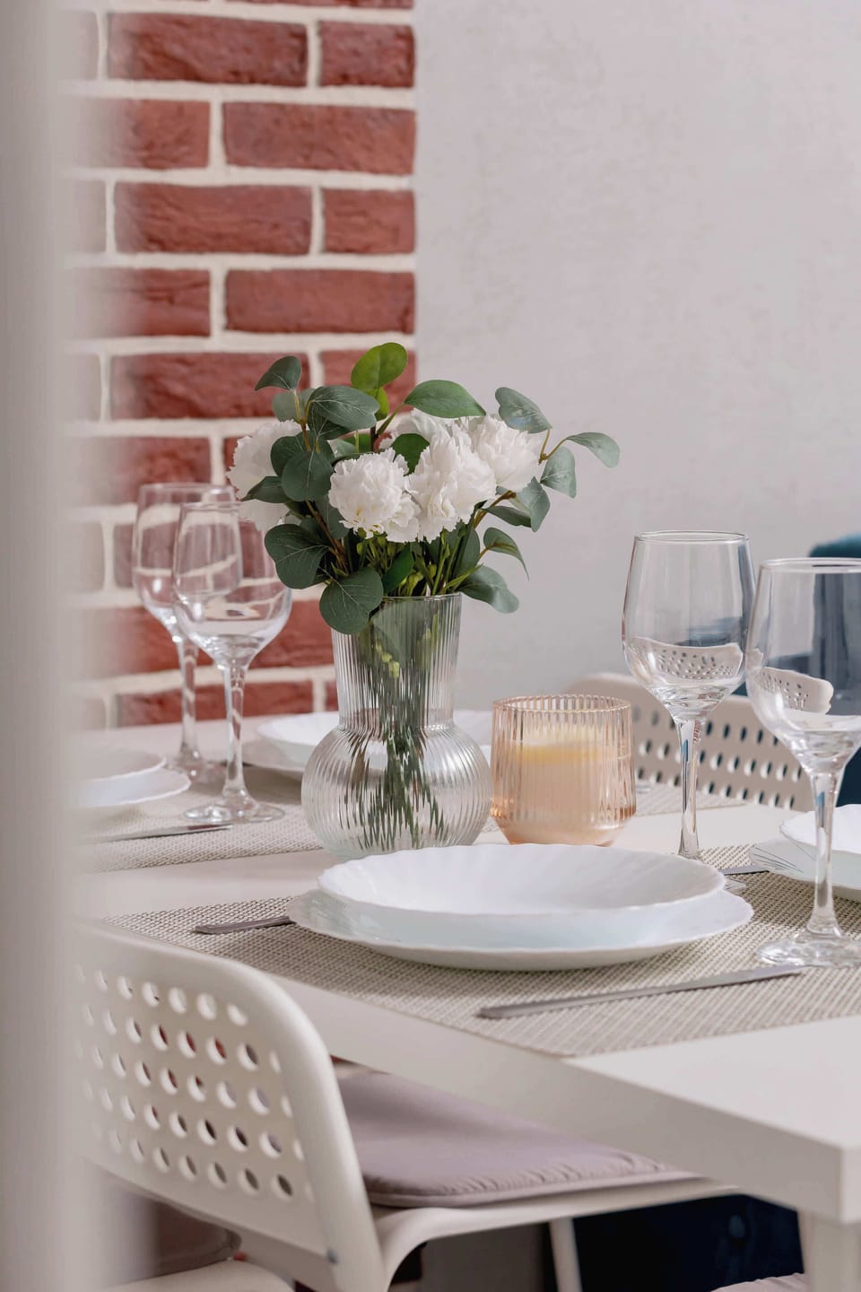 A beautifully set dining table with plates, glasses, and a floral centerpiece. The brick wall adds a warm touch.