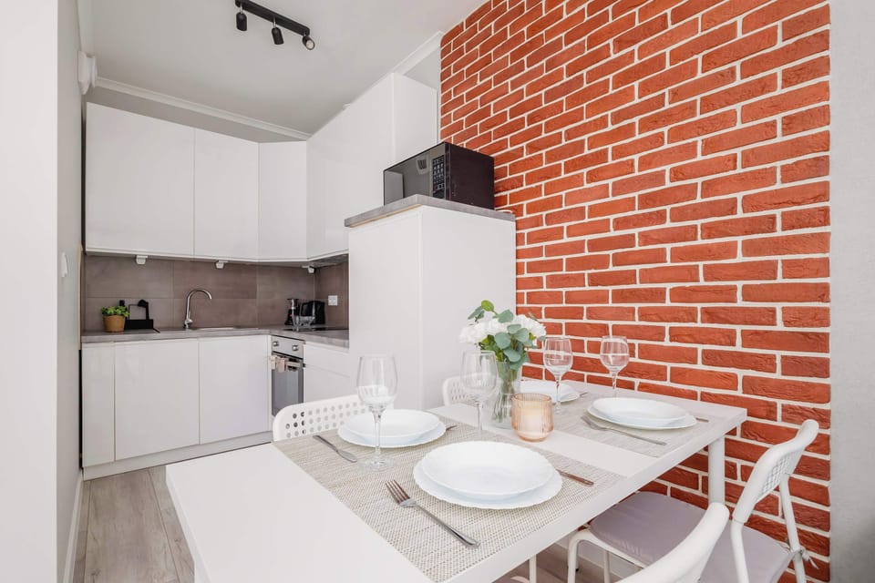 A dining area with a table set for a meal, next to a modern kitchen. The combination of brick and white cabinetry creates a warm and contemporary feel.