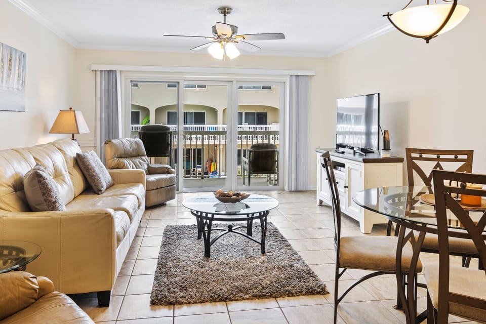 Bright beachside living area with plush seating, flat-screen TV, and dining space. Sliding doors open to a private balcony, perfect for enjoying coastal breezes and poolside views.