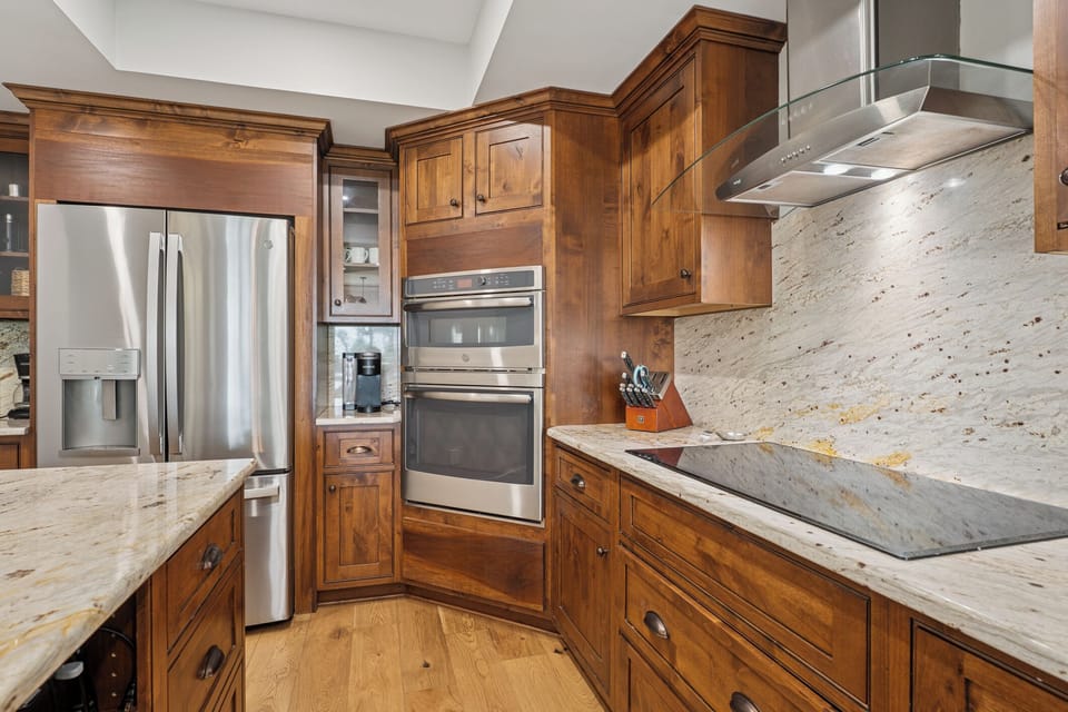 This well-appointed kitchen features rich wooden cabinetry, light-colored granite countertops, a built-in oven, and a large stainless steel refrigerator