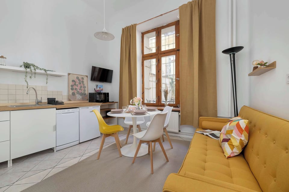 A modern open kitchen with white cabinets and appliances, leading into a bright dining area with a yellow sofa.
