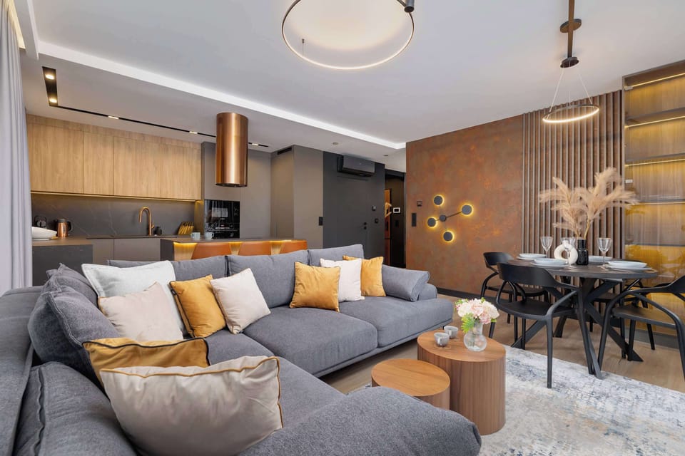A modern living room with a cozy gray sofa, stylish coffee table, and a large rug. The decor includes golden accents and a ceiling-mounted round chandelier. In the background, a dining table with chairs and the open-plan kitchen can be seen.