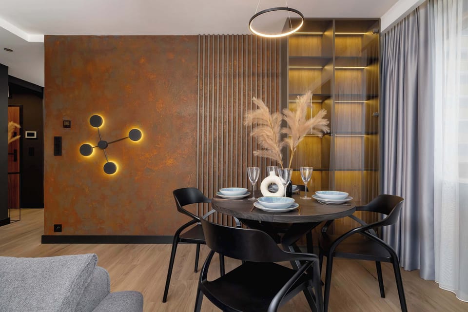 The dining area features a round table with four chairs in soft gray fabric. Stylish decorative pieces are placed on the table, and the kitchen is visible in the background.

