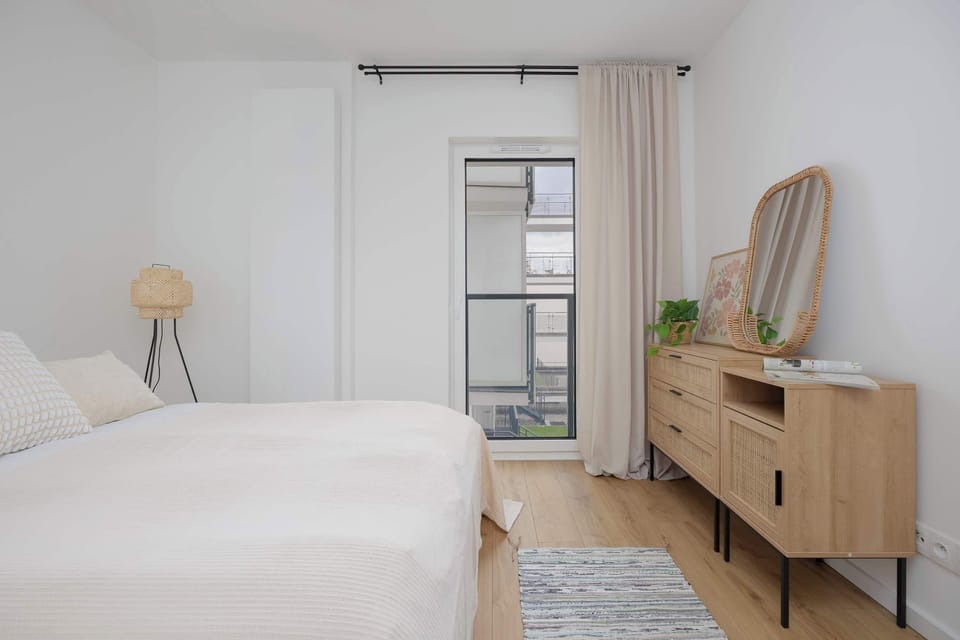 A larger view of the same bedroom showing a wooden dresser, large windows, and a serene, airy atmosphere.

