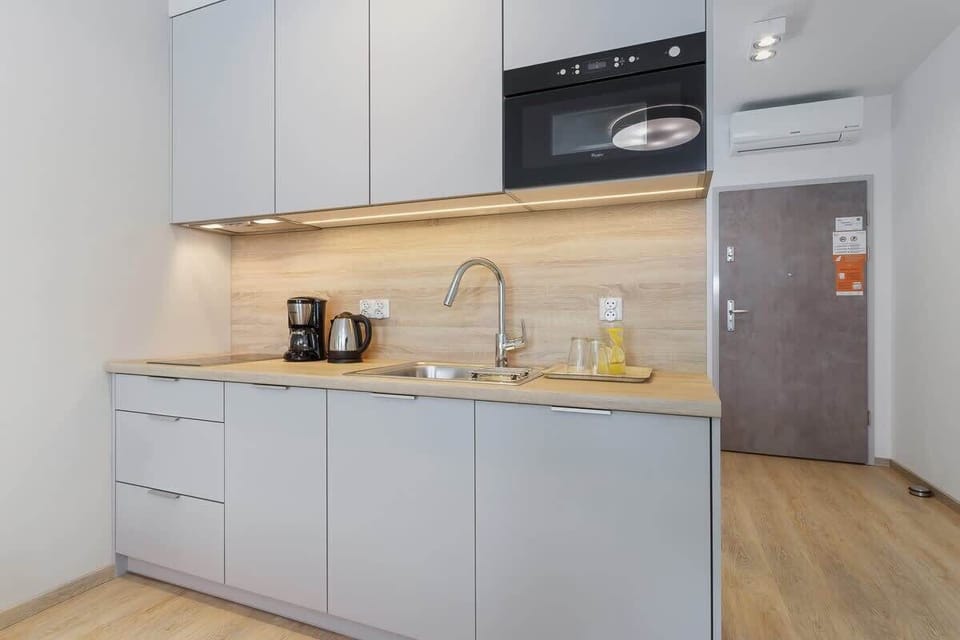 A compact kitchenette with light gray cabinets, wood countertops, and essential appliances. The open shelving adds to the minimalist aesthetic.