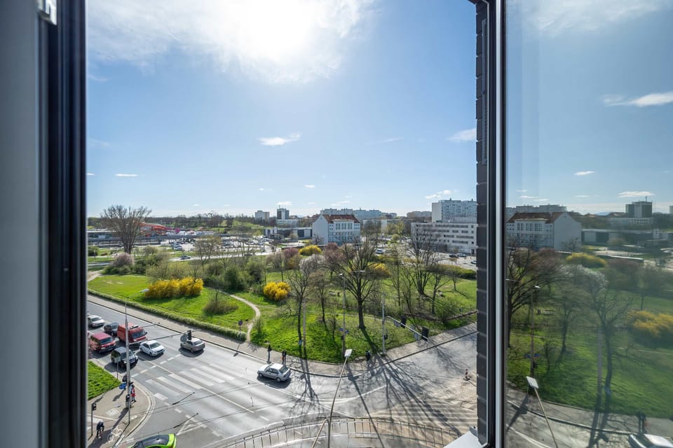 A panoramic view from the window showing a vibrant green park area and residential buildings under a clear blue sky. The view adds a refreshing contrast to the minimalist indoor design.