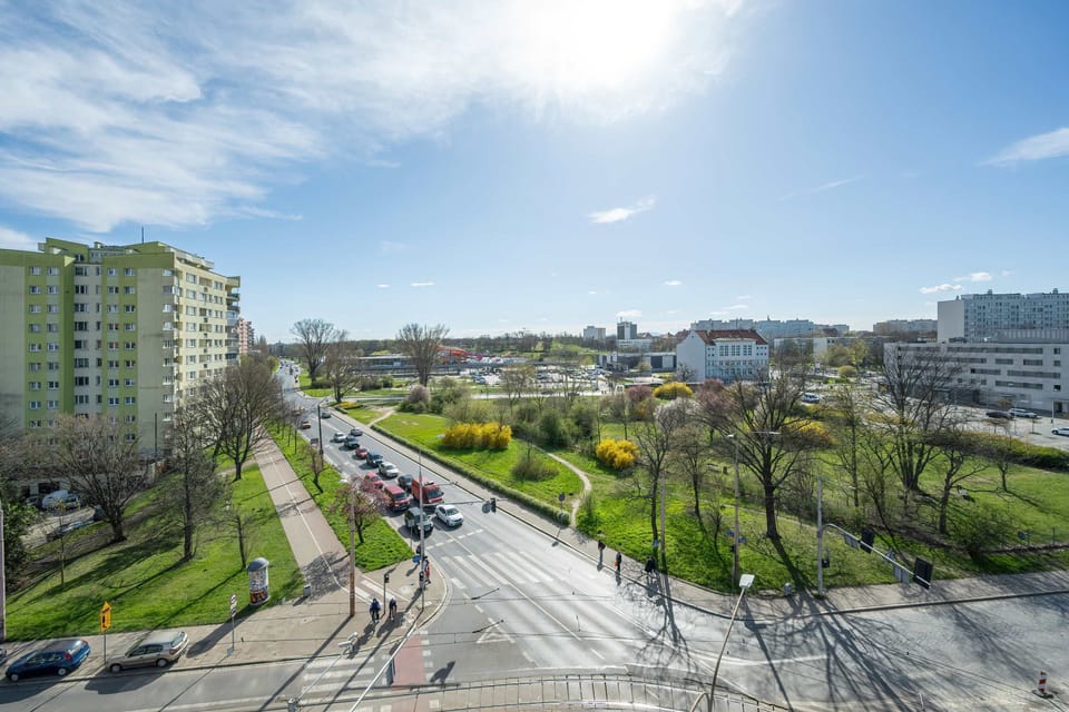 A scenic view of a green urban landscape seen from the apartment’s window, with trees and residential buildings visible in the distance. The sunny day adds to the vibrant, fresh atmosphere.