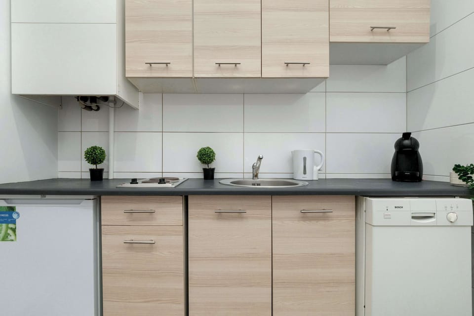 A compact kitchenette with light wood cabinetry, white tiled walls, and modern black countertops.