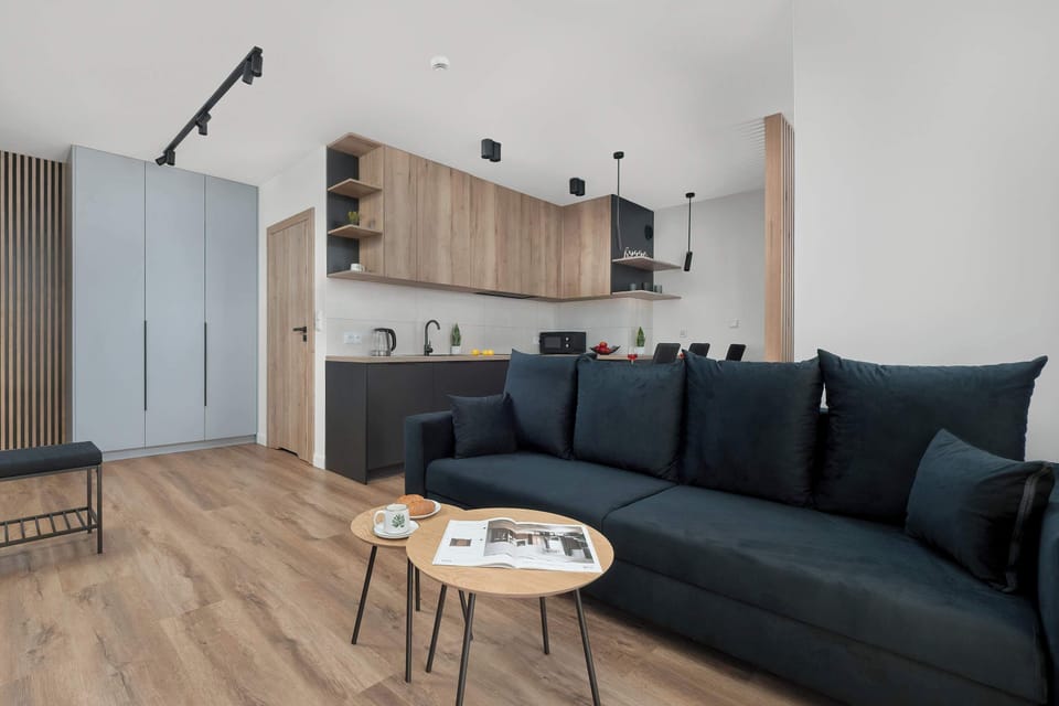 Bright living area with a dark grey sofa, light wooden flooring, and large windows covered with light curtains.