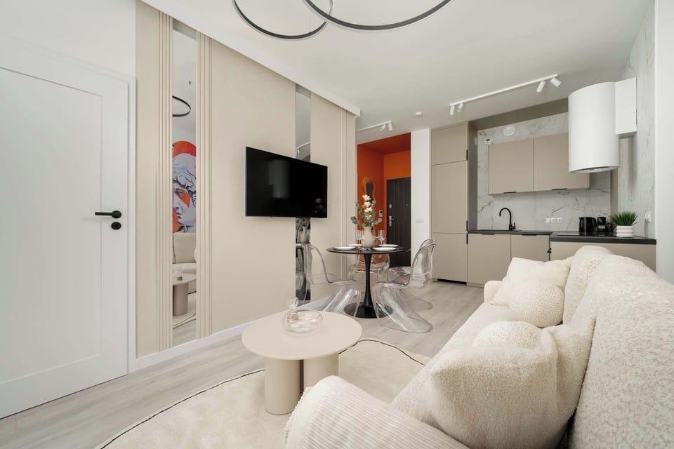 Bright and cozy living room with beige sofa, TV, round table and kitchenette.