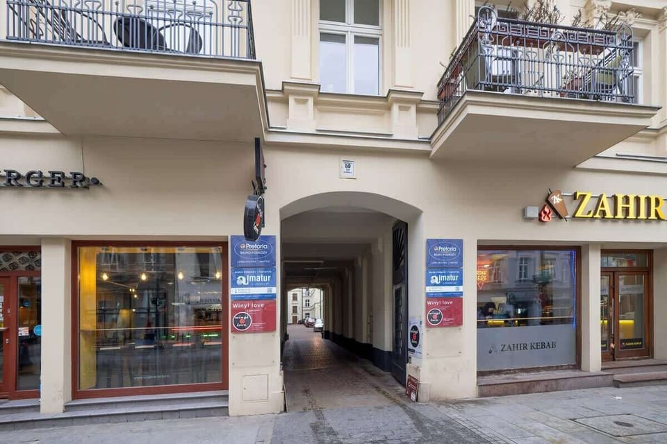 Entrance to a residential passageway, flanked by a Żabka store and another shop. The archway leads into a courtyard or hallway.
