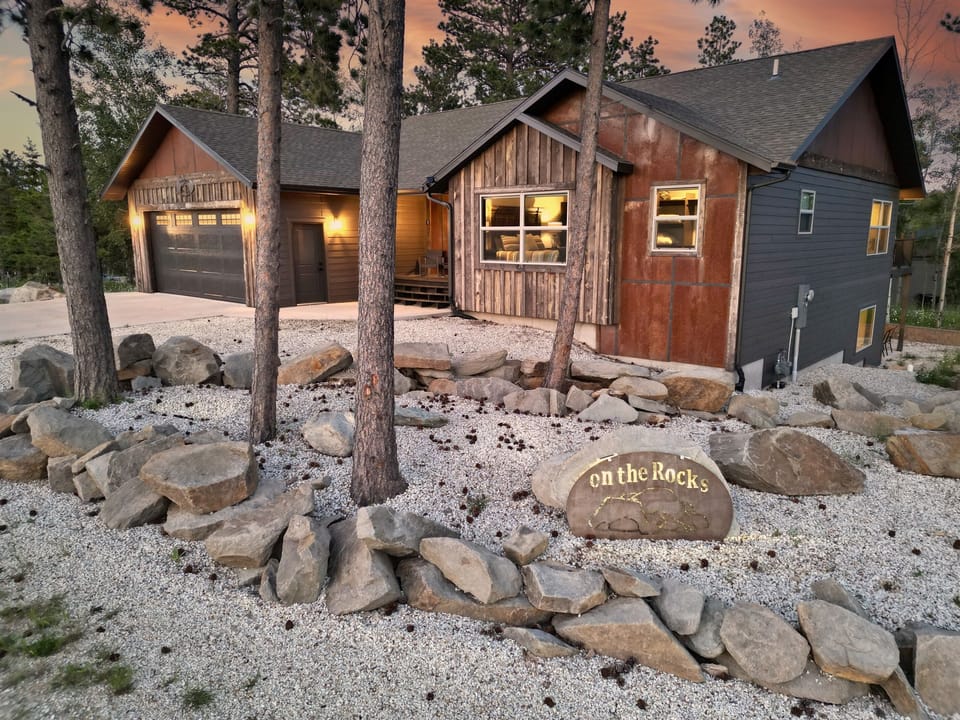 Imagine arriving at "On The Rocks" in Powder House Pass, stepping into the rustic charm of this inviting cabin. Picture the warm lights glowing against the evening sky, the crunch of gravel, and the natural rock garden welcoming you to a peaceful retreat.