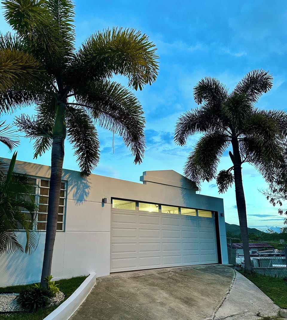 Modern tropical-style home with bright white exteriors, palm trees, and a private driveway. A relaxing escape just minutes from the Coamo Hot Springs, perfect for guests seeking comfort, style, and a touch of Caribbean sunshine.